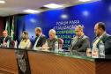 Foto lateral da mesa de autoridades do Fórum da Pessoa com Deficiência. Compõe a mesa, quatro homens duas mulheres. Os homens estão de blazer cinzas e azuis. 