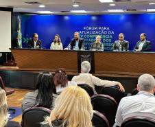 Foto ampla, com várias pessoas aparecendo de costas. Em frente à elas estão outras sentadas à mesa de abertura do Fórum da Pessoa com Deficiência. 