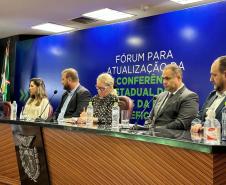 Foto lateral da mesa de autoridades do Fórum da Pessoa com Deficiência. Compõe a mesa, quatro homens duas mulheres. Os homens estão de blazer cinzas e azuis. 
