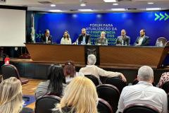 Foto ampla, com várias pessoas aparecendo de costas. Em frente à elas estão outras sentadas à mesa de abertura do Fórum da Pessoa com Deficiência. 