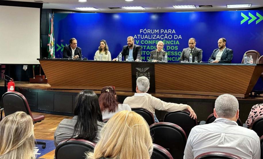 Foto ampla, com várias pessoas aparecendo de costas. Em frente à elas estão outras sentadas à mesa de abertura do Fórum da Pessoa com Deficiência.