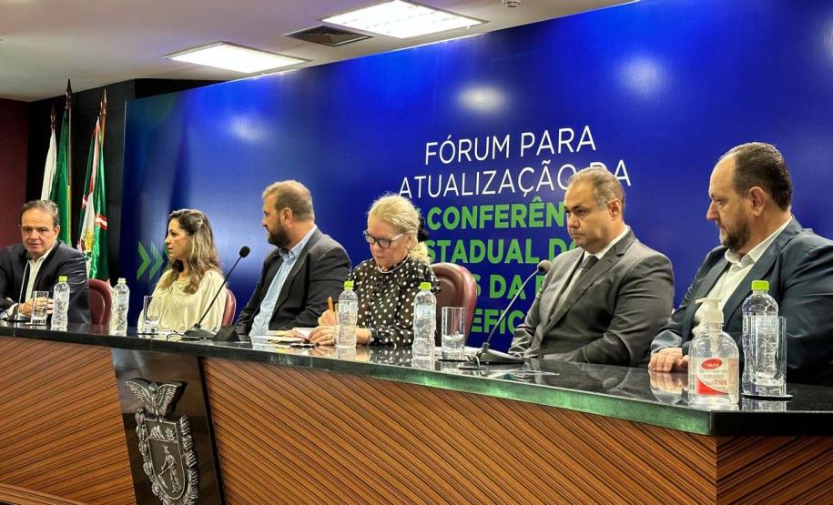 Foto lateral da mesa de autoridades do Fórum da Pessoa com Deficiência. Compõe a mesa, quatro homens duas mulheres. Os homens estão de blazer cinzas e azuis.